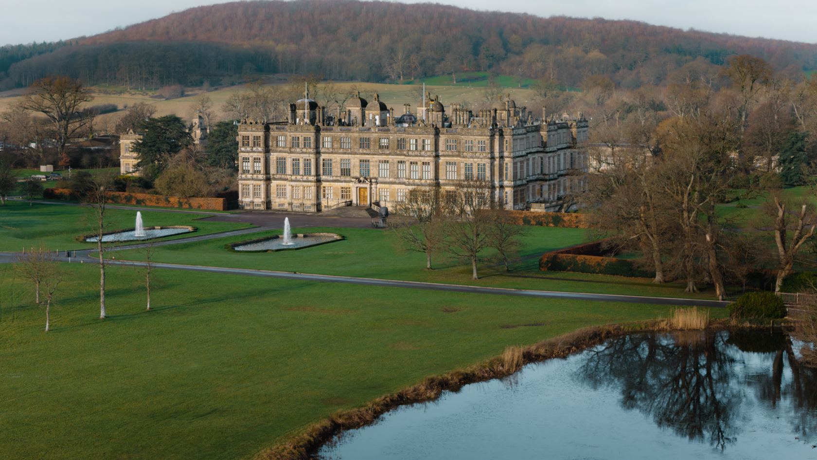 A drone shot of Longleat house in the middle distance with a lake in the foreground and a wooded hill in the background