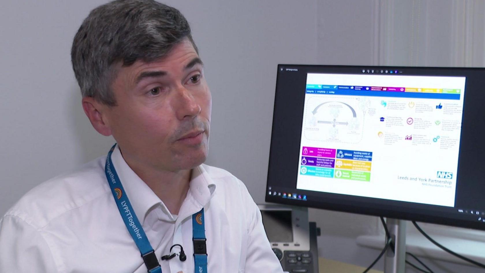 Dr Chris Hosker, wearing a white shirt and NHS lanyard, in front of a computer screen