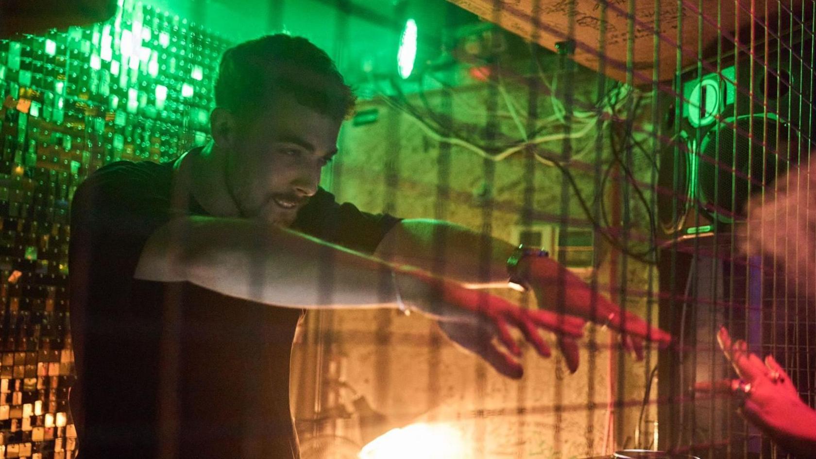 A male DJ dances to music with his arms lifted out in front of him and pointing towards people in a crowd. 