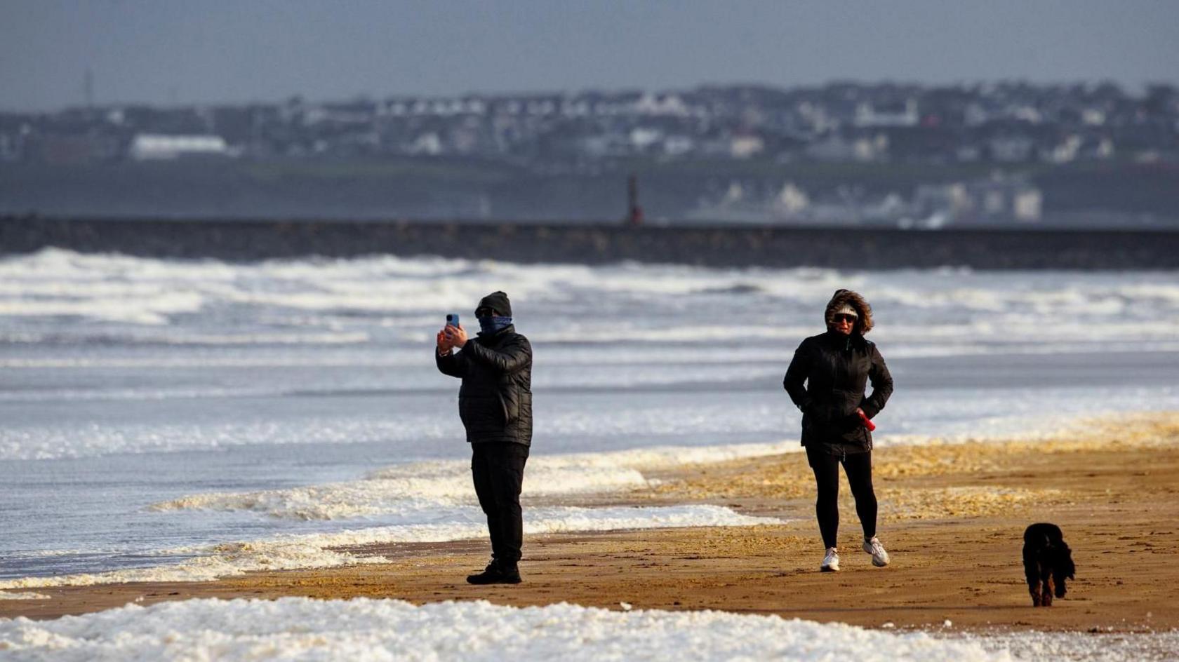 People wrapped in jackets and hats walk along a beach. 