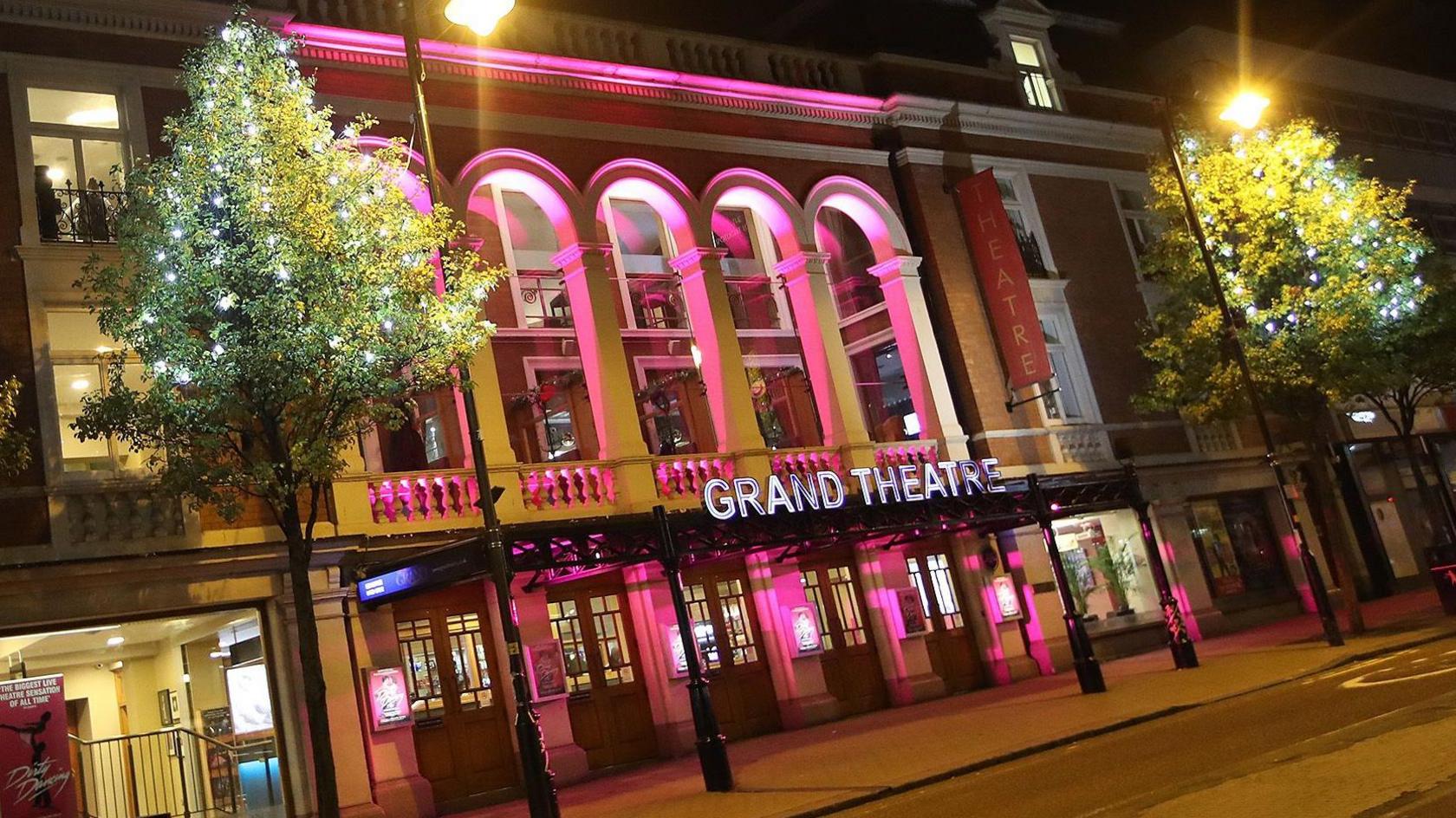 The front of the Grand Theatre lit up pink at night, there are two tries on either side with lights in the branches