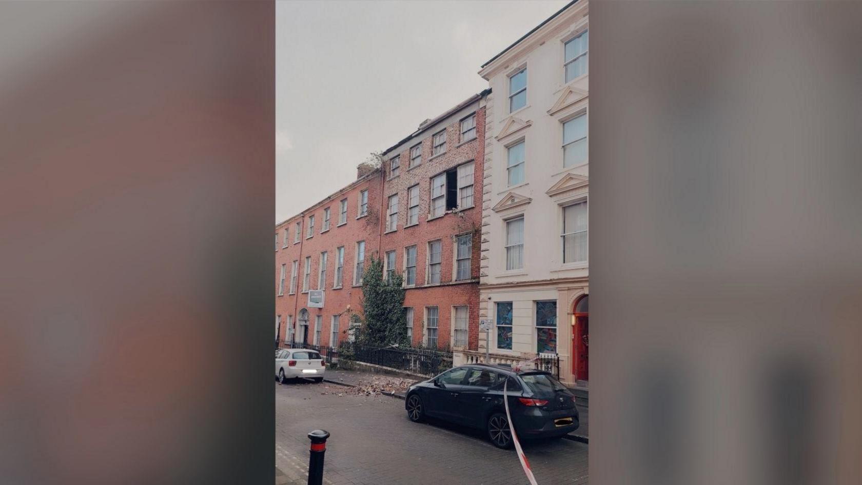 A wider view of terraced buildings on Pump Street in Derry. There is a gap between two second-storey windows at the front of a red-bricked building in the middle of the photo. There is a pile of red bricks on the pavement below.