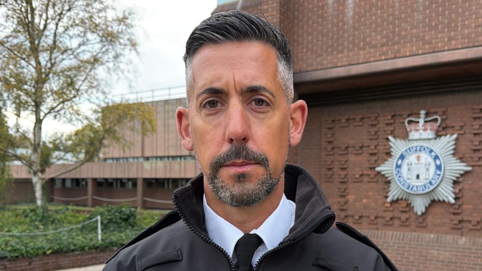 Ch Insp Gary Miller is stood in the middle of the image looking at the camera. He has short grey hair and is wearing a white shirt, a black tie and black coat.