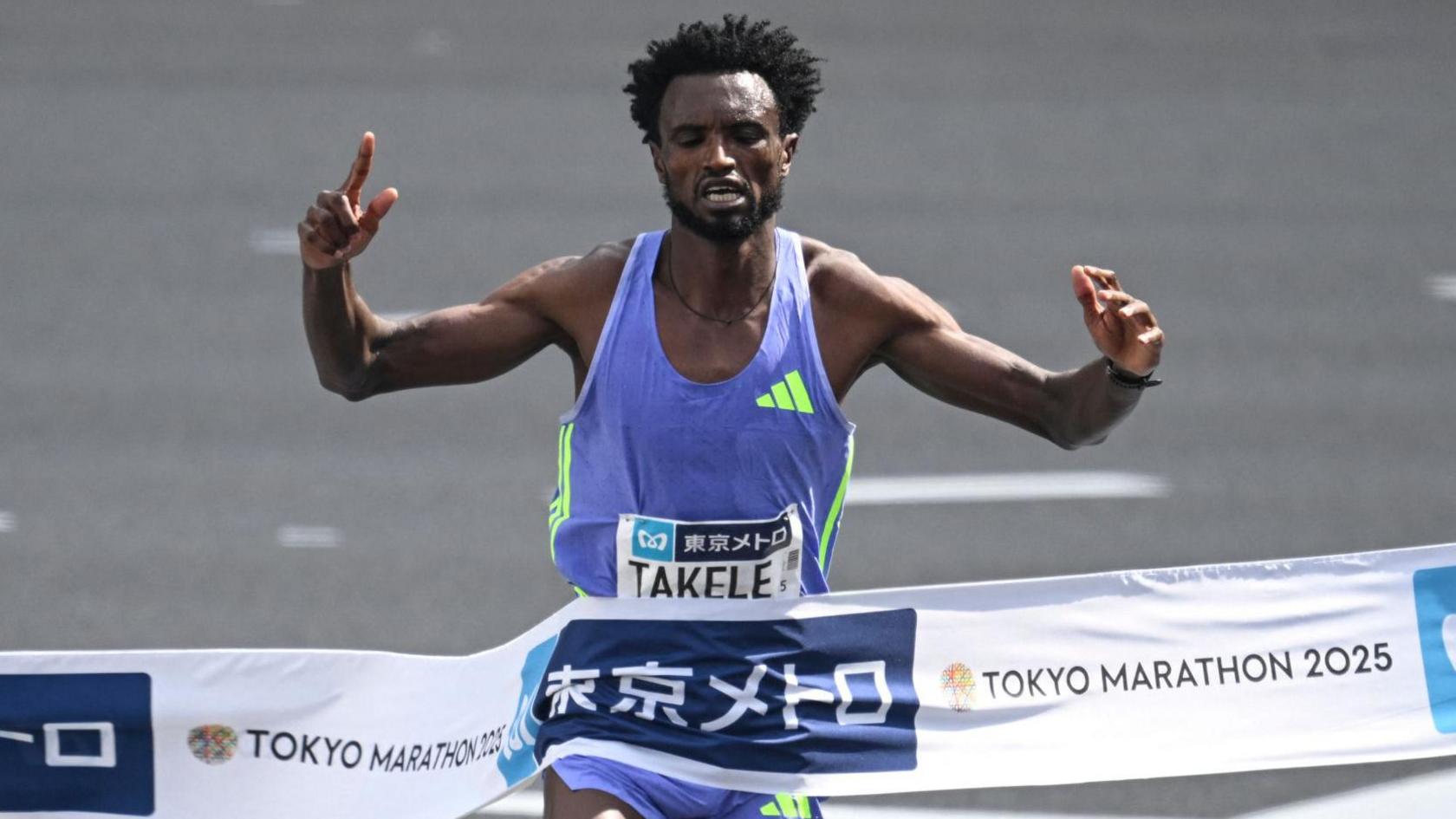 Tadese Takele, wearing purple running top and shorts, holds his hands in the air as he breaks a finish line tape which reads Tokyo Marathon 2025 and also features Japanese text symbols