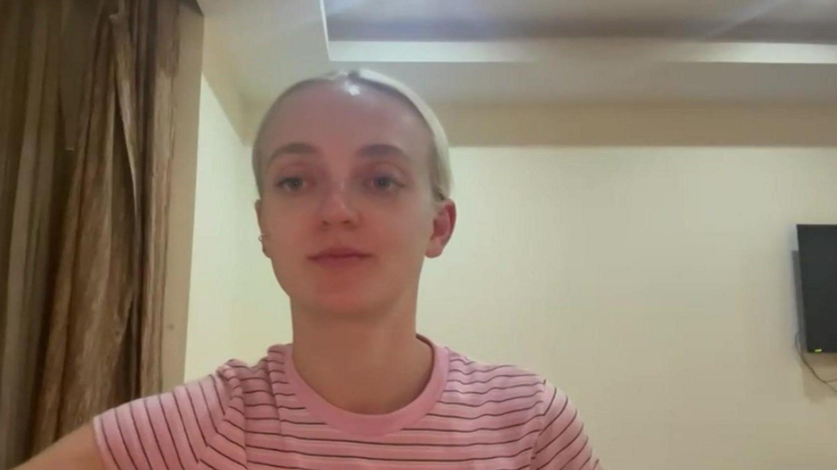 Poppy Cleary vesh një bluzë me vija rozë dhe të zeza ndërsa shikon drejtpërdrejt nga kamera. Ajo duket sikur është në një dhomë hoteli me mure ngjyrë krem ​​dhe perde kafe mbi shpatulla.