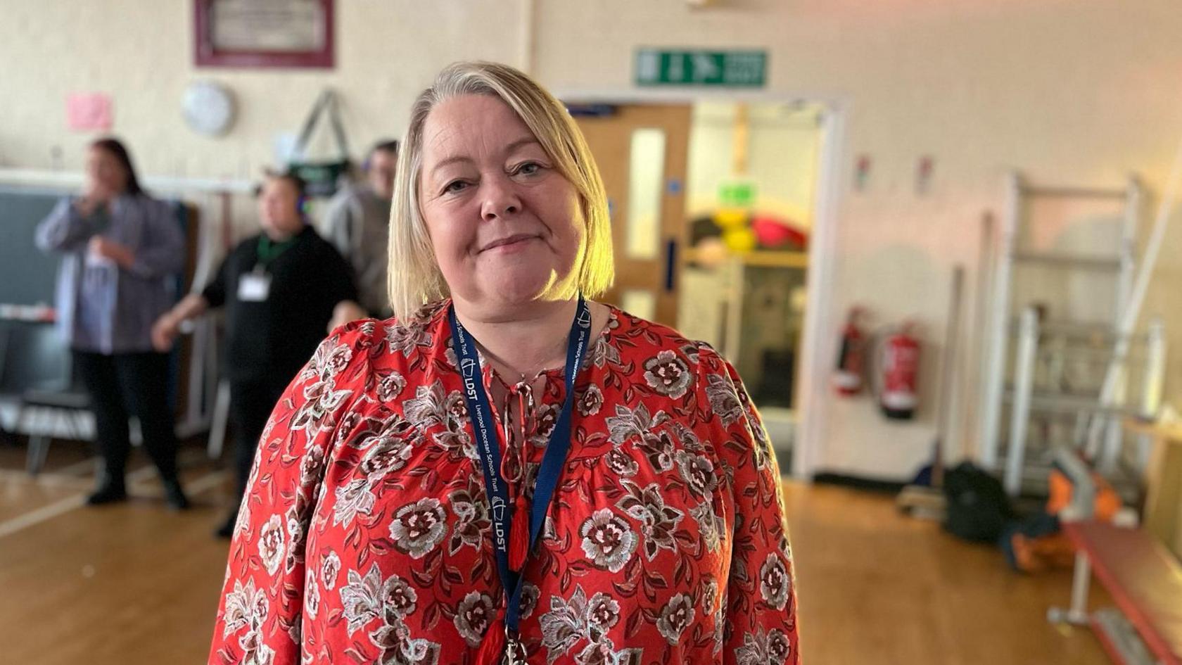 Linda Thompson smiles at the camera as she stands in the school hall. She has blonde bobbed hair and is wearing a red patterned top. Three adults can be seen blurred out in the background.