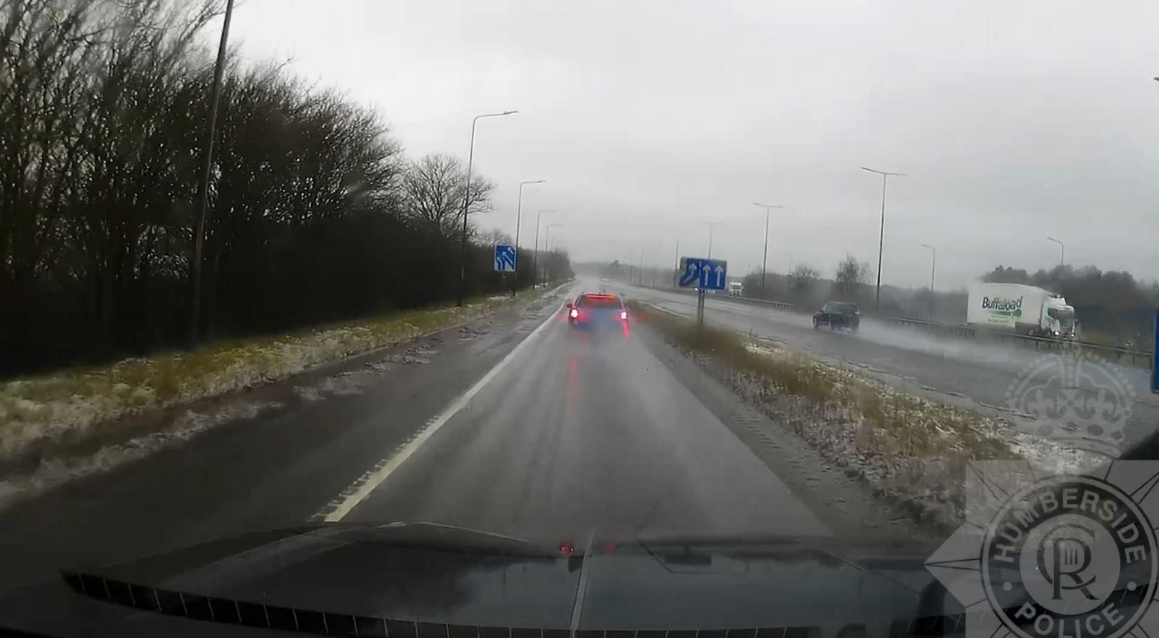 Slipway onto a motorway. A blue car is in front and on the left is a black car. The weather is wet, clouds are grey and gloomy.