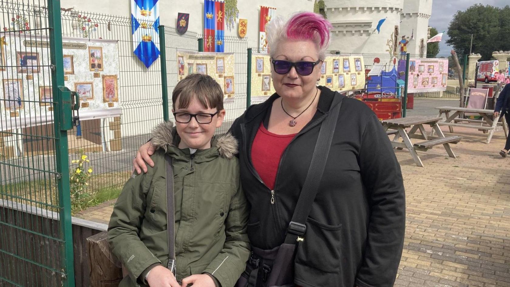 Saul stands beside his mum at the exhibition. She has her arm around him. Saul has brown hair and wears glasses, wearing a green parka. Sue as grey and pink hair, wears a red top with a black jacket that is partially zipped up.