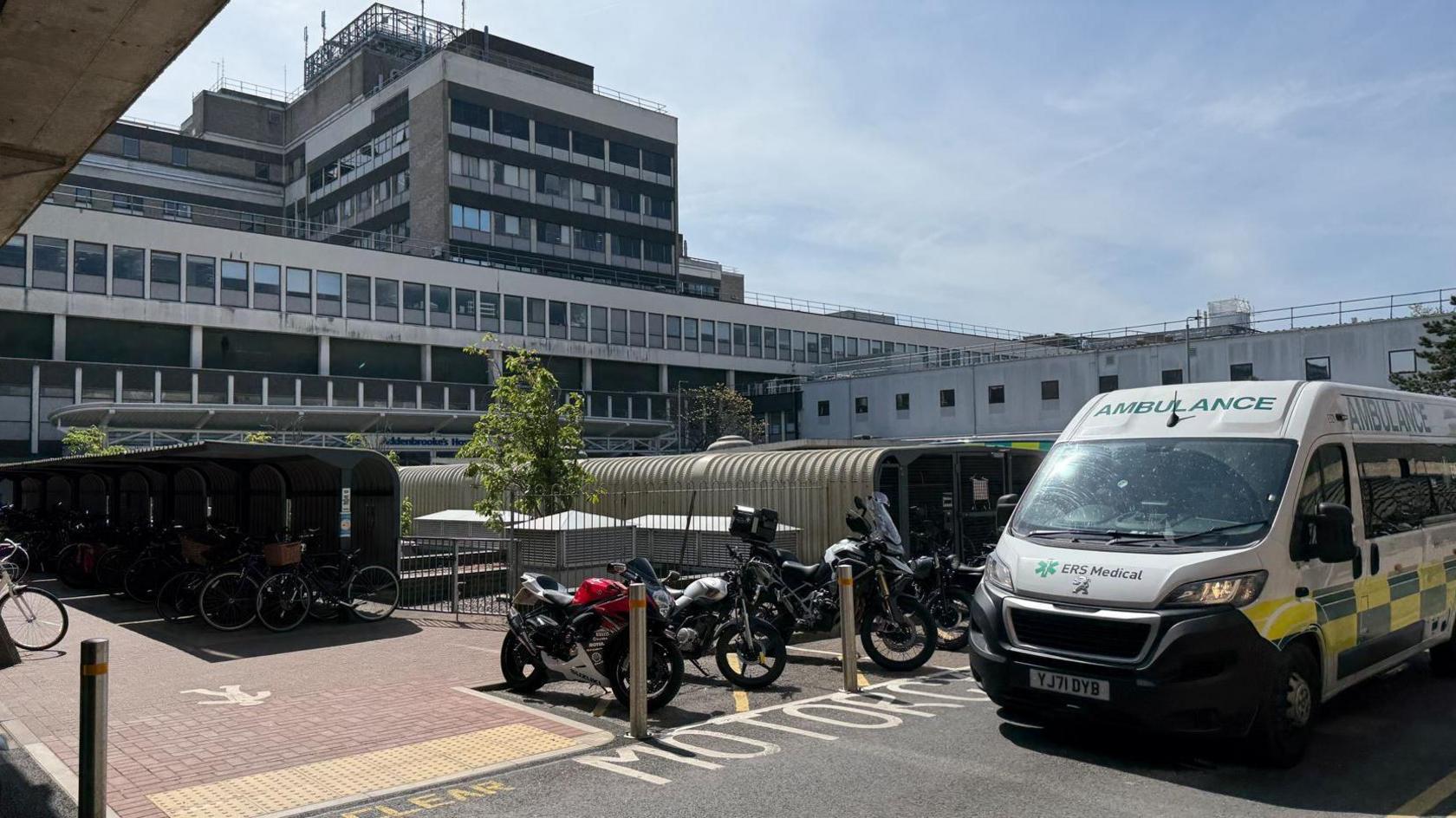 The exterior of Addenbrooke's Hospital, a white and grey building, with a parked ambulance and motorcycles in the foreground
