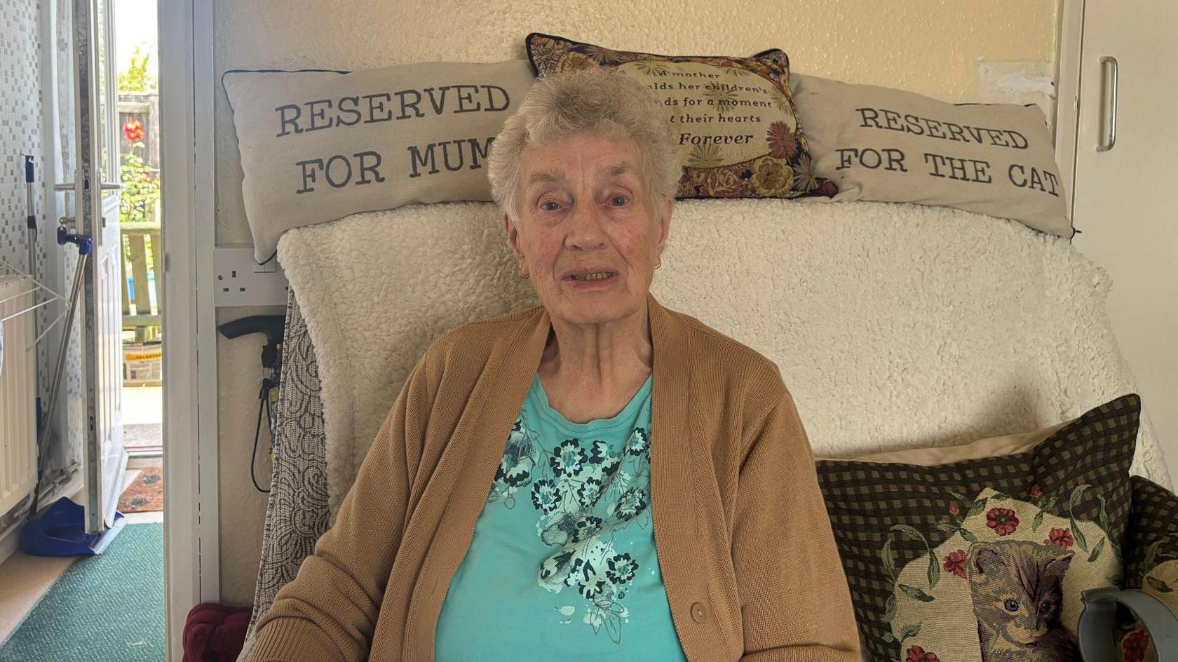 An older woman with short grey hair sitting in a chair. She is wearing a brown cardigan and blue top.