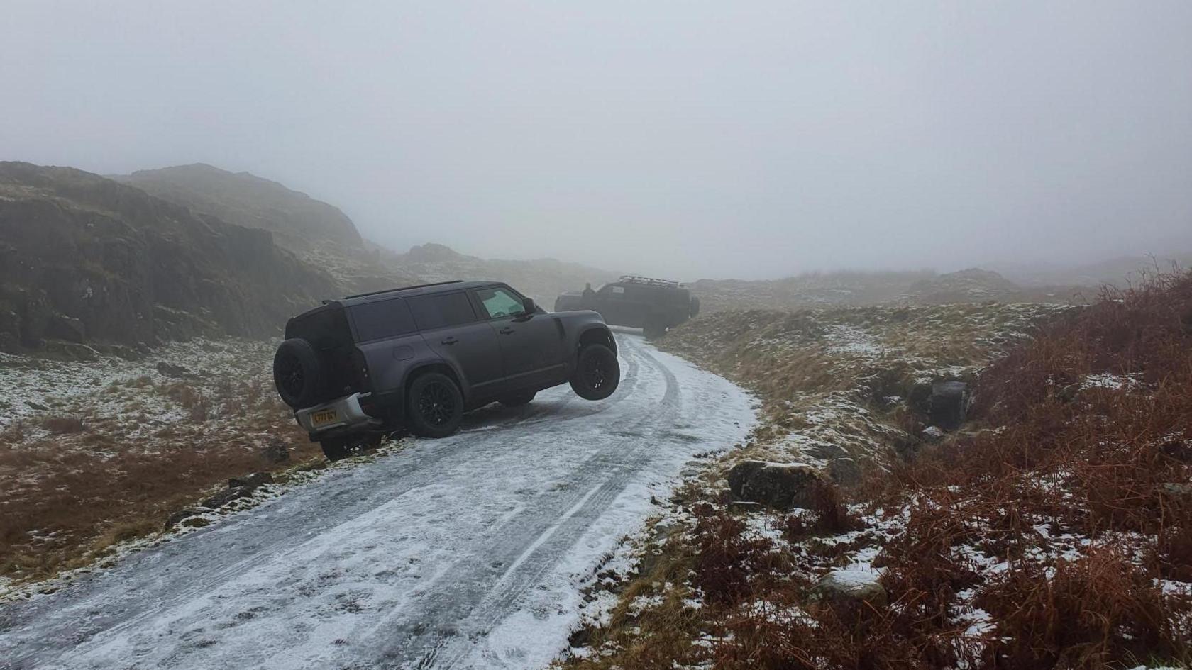 An icy single track road with two black cars run off it. The road is bordered by tall mountains.