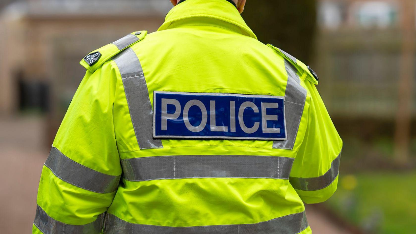 A police officer seen on a residential street from behind. He is wearing a high-vis jacket with the words "police" on