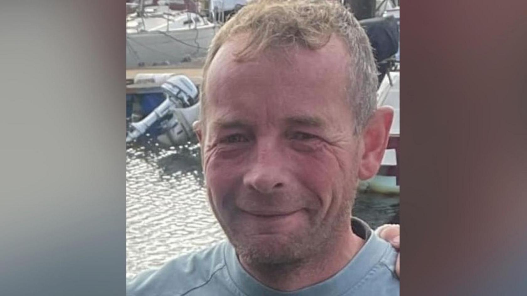 Lee Scott, who has light brown short cropped hair and stubble on his face, and is wearing a light blue T-shirt. There are water and boats behind him.