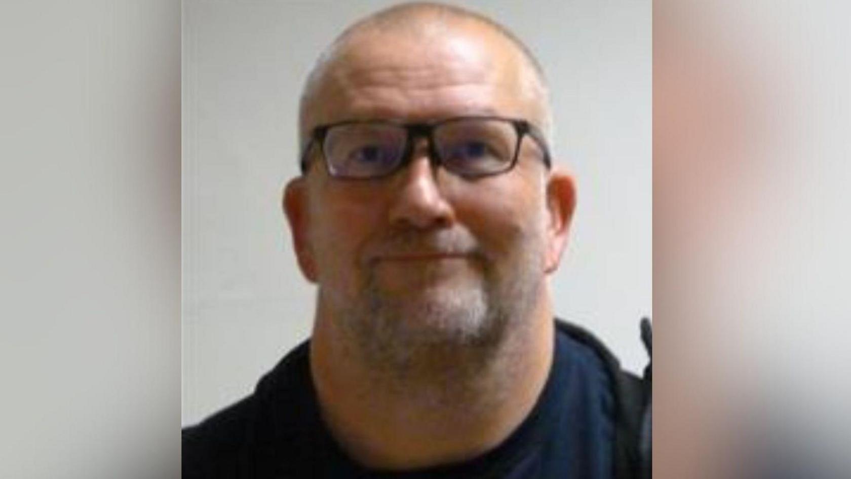 White man in a blue T-shirt looking at the camera. He has slight grey subtle. He's wearing glasses with a dark coloured frame.