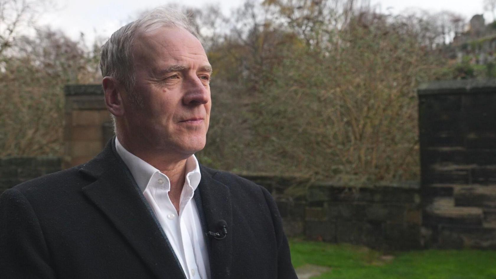 Angus Farquhar, a white-haired man in a black blazer and white shirt looks to the left in the churchyard of Glasgow Cathedral, grass and trees and a stone wall behind him.
