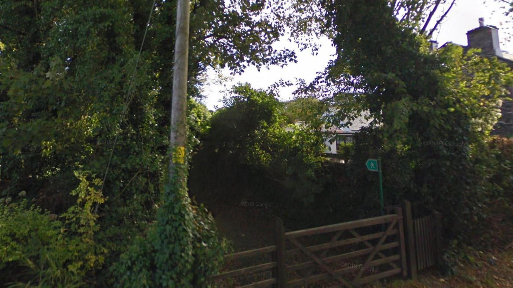The entrance to the footpath, a fence is in place and there are lots of trees around.