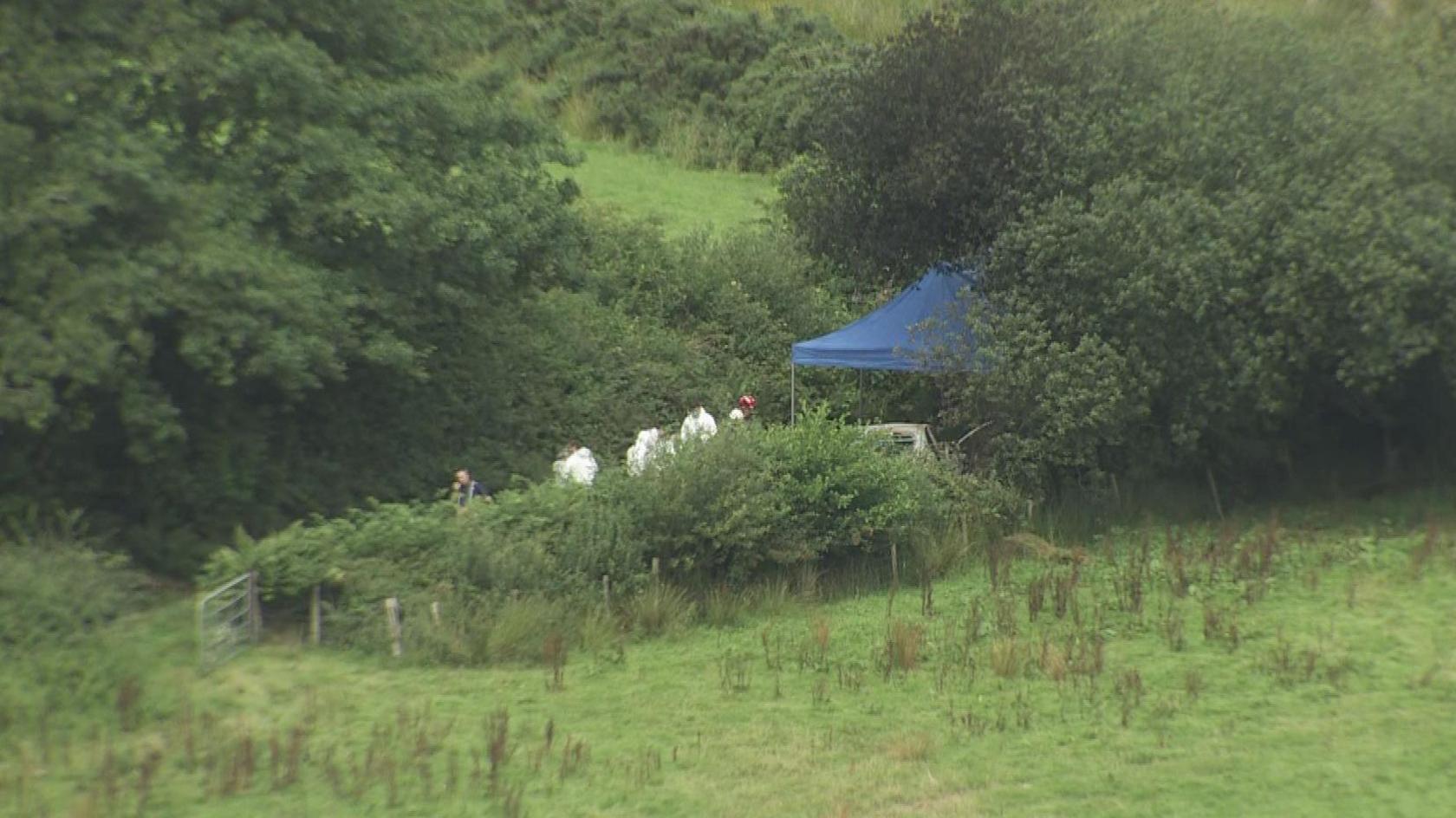 A blue tent has been erected on a country road. The image is taken from a distance, but you can see a number of people dressed in all-white forensic suits. They are surrounded by trees and grass. 