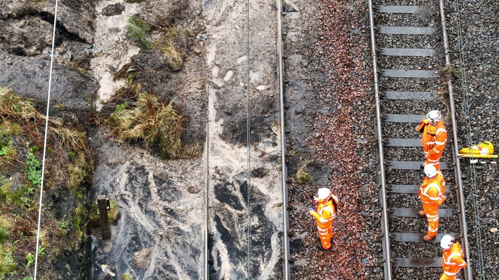 Four workmen in orange hi-vis stand on a railway track as they look at the side of it which appears to show a muddy ditch and debris across one of the tracks.