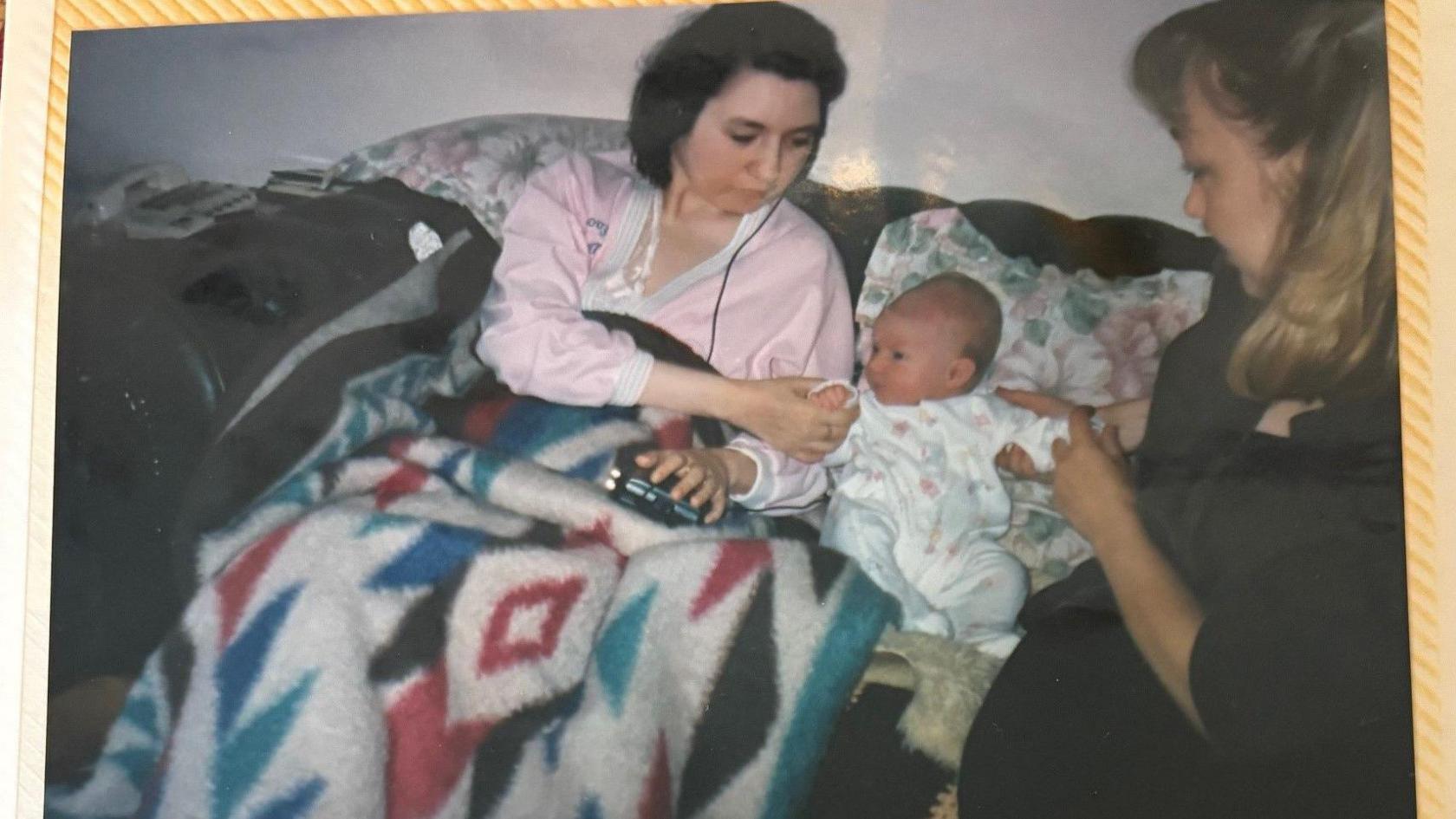 A photo on a page of a woman with shoulder-length brown hair, in a pink nightie under a multi-colour blanket, sat on a sofa next to a baby.