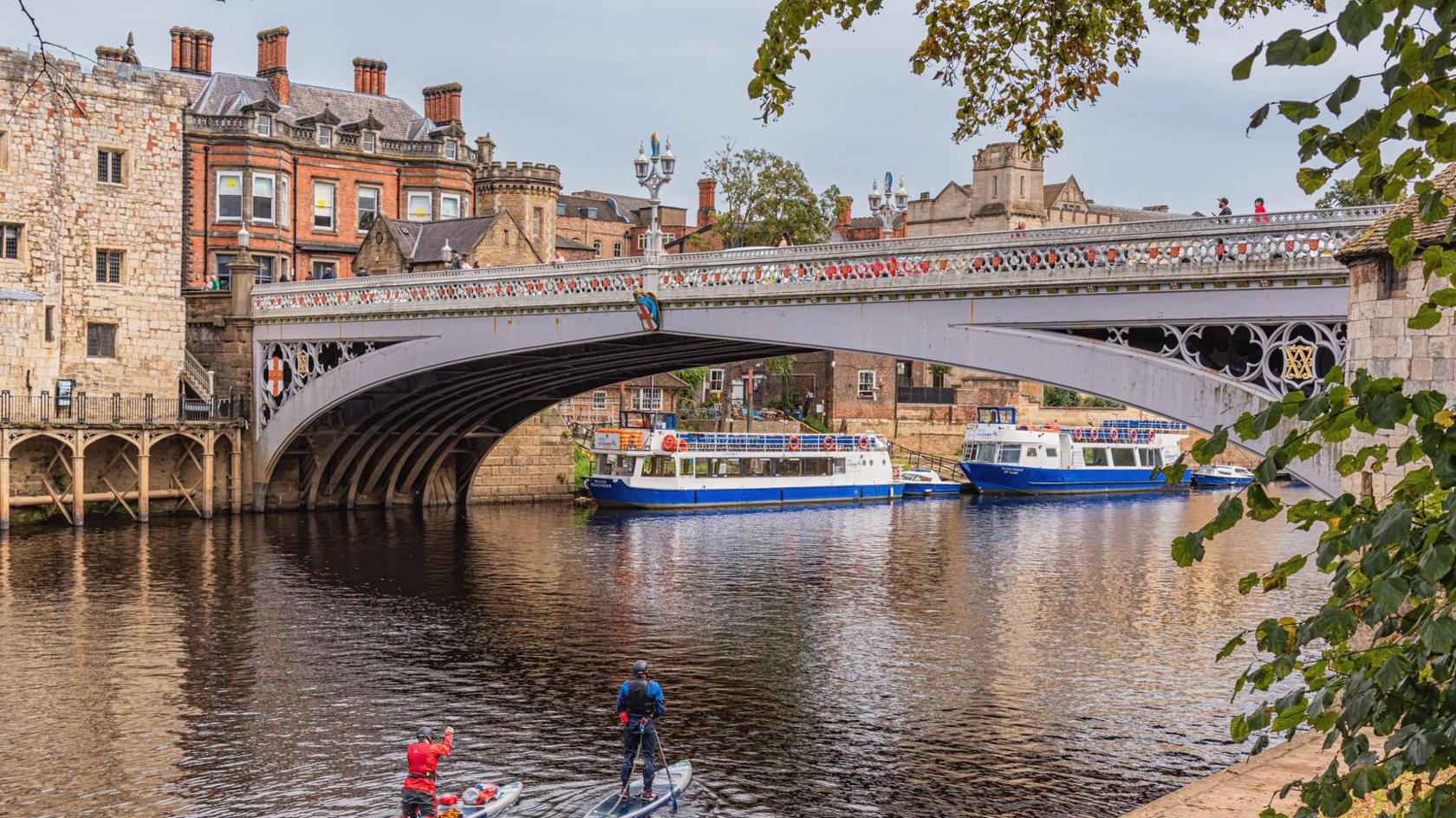 A view of the River Ouse in York with the Victorian Lendal Bridge, an iron structure in white with gilding and red coat of arms on its arches, through the bridge two river cruisers in white and blue can be seen