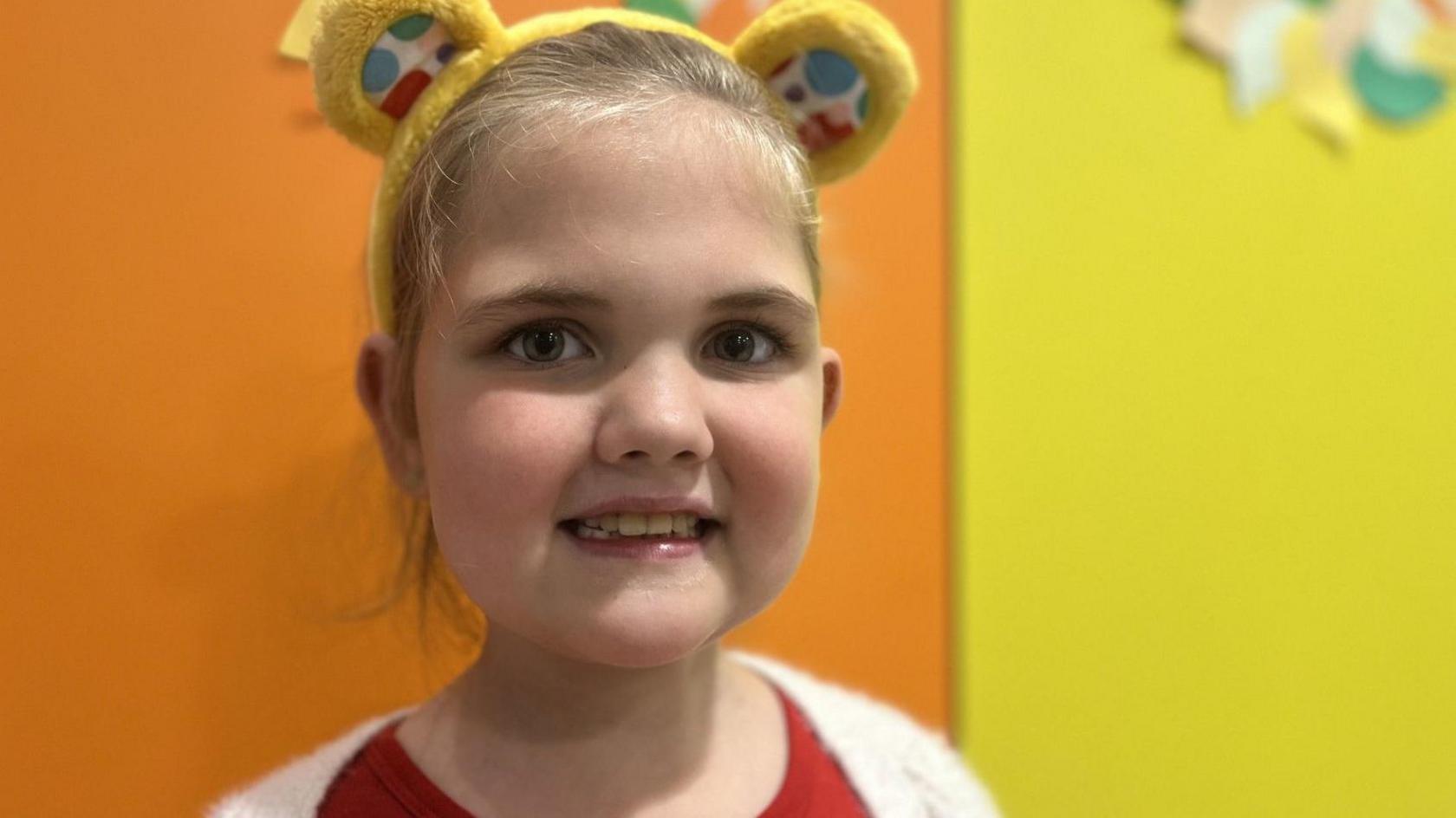 A little girl smiles straight at the camera. She has blonde hair tied back and is wearing a yellow fluffy headband with teddy-bear style "Pudsey" ears. She has dark brown eyes. She is wearing a red top and white cardigan. The background is bright and colourful with blocks of orange and yellow.