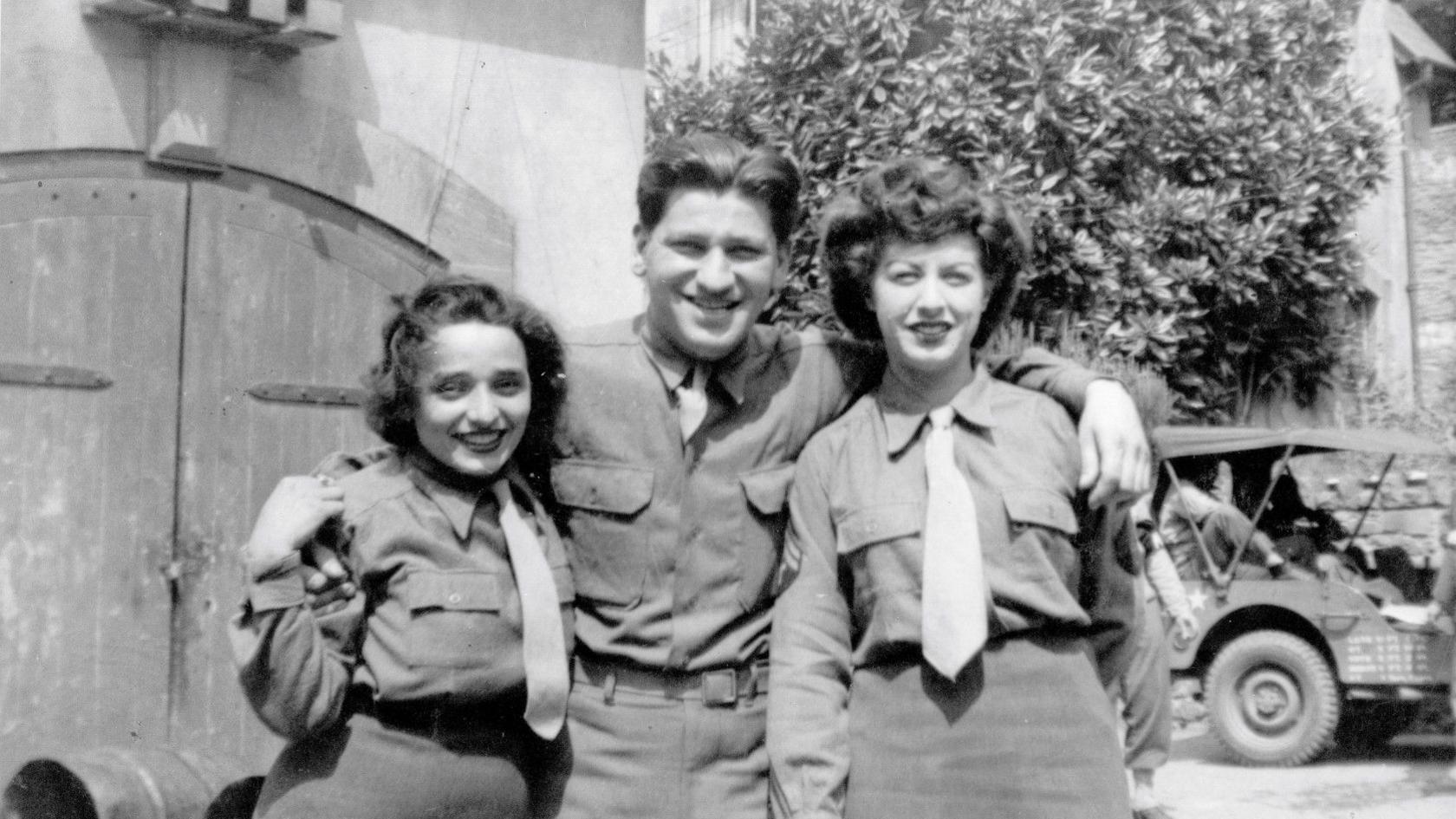 A black and white image of a man in a shirt, with his arms around two women, who are both in a uniform comprising shirts, ties and skirts. They stand in front of stable doors, with a buggy-style vehicle in the background.