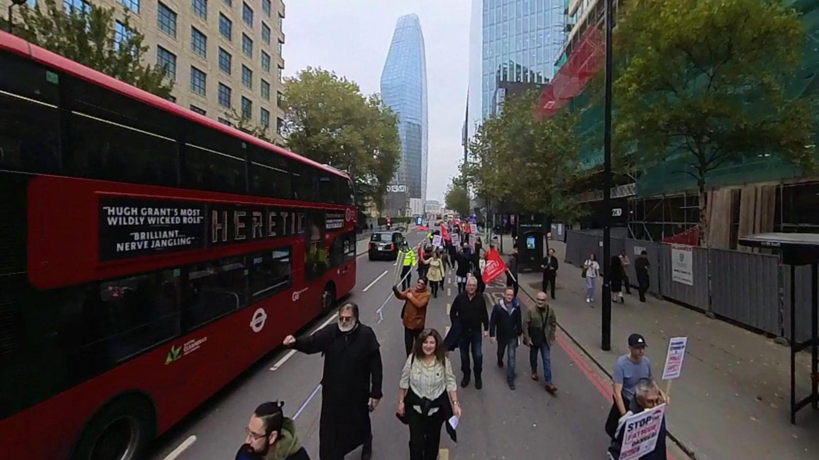 Image of people walking along a road as a bus passes by. Some people are holding placards.