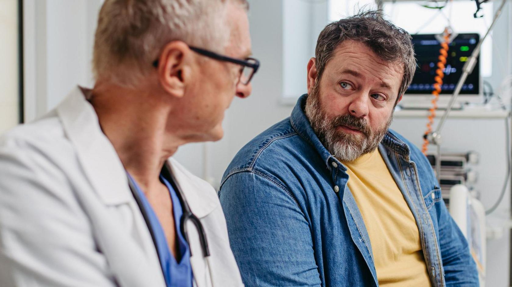A doctor wearing scrubs and a white coat talks to a middle-aged man wearing a yellow t-shirt and denim shirt.