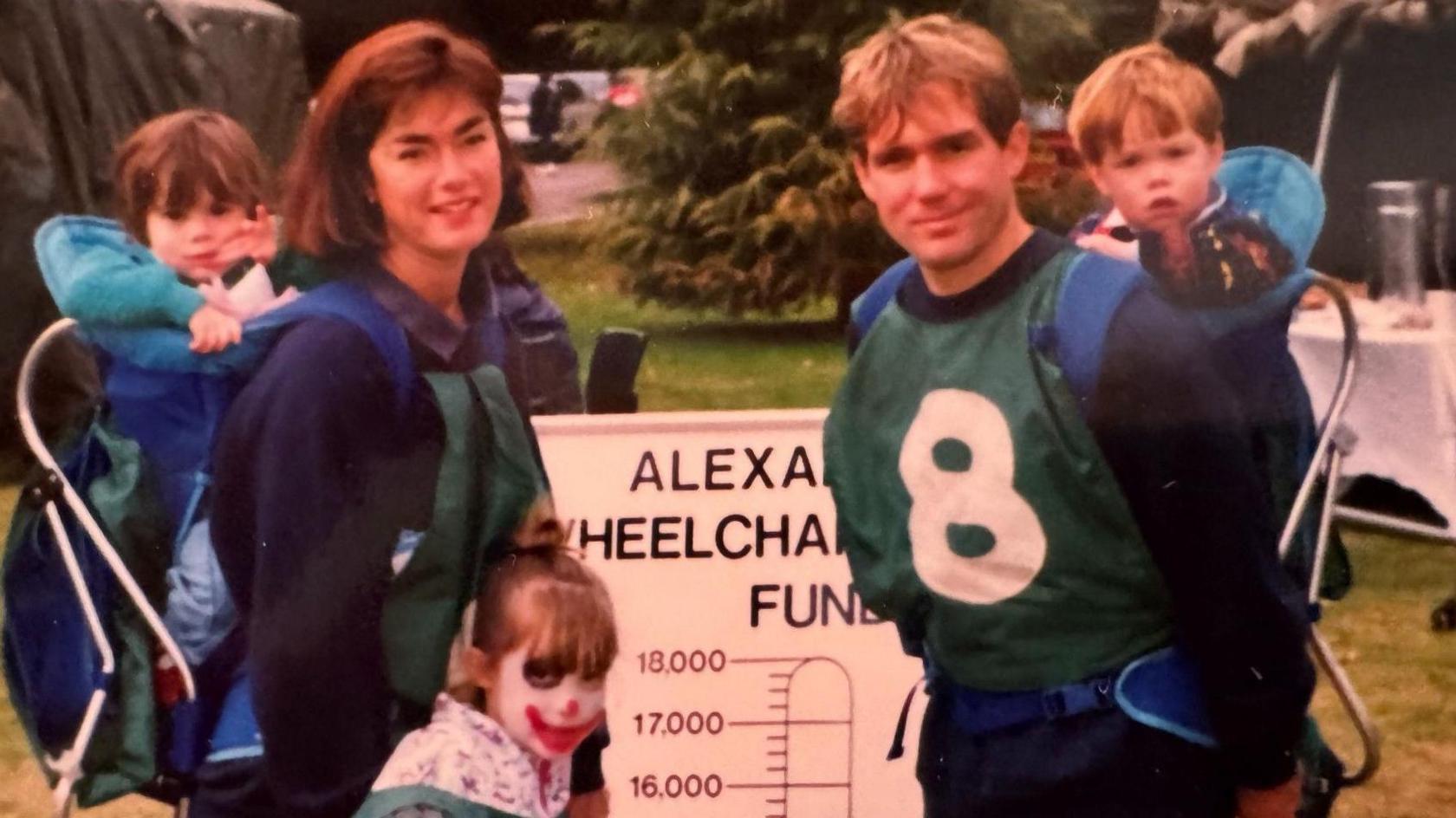 Maj Sparks and his wife, Sue, carrying a toddler each in a backpack with another child standing between them. She has clown face paint on.