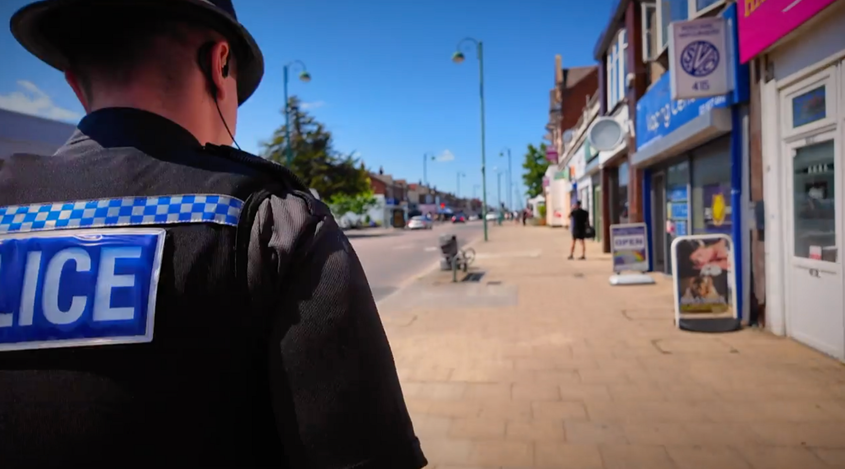 A police officer in uniform walks down a High Street pavement lined with shops, set against a blue sky. 