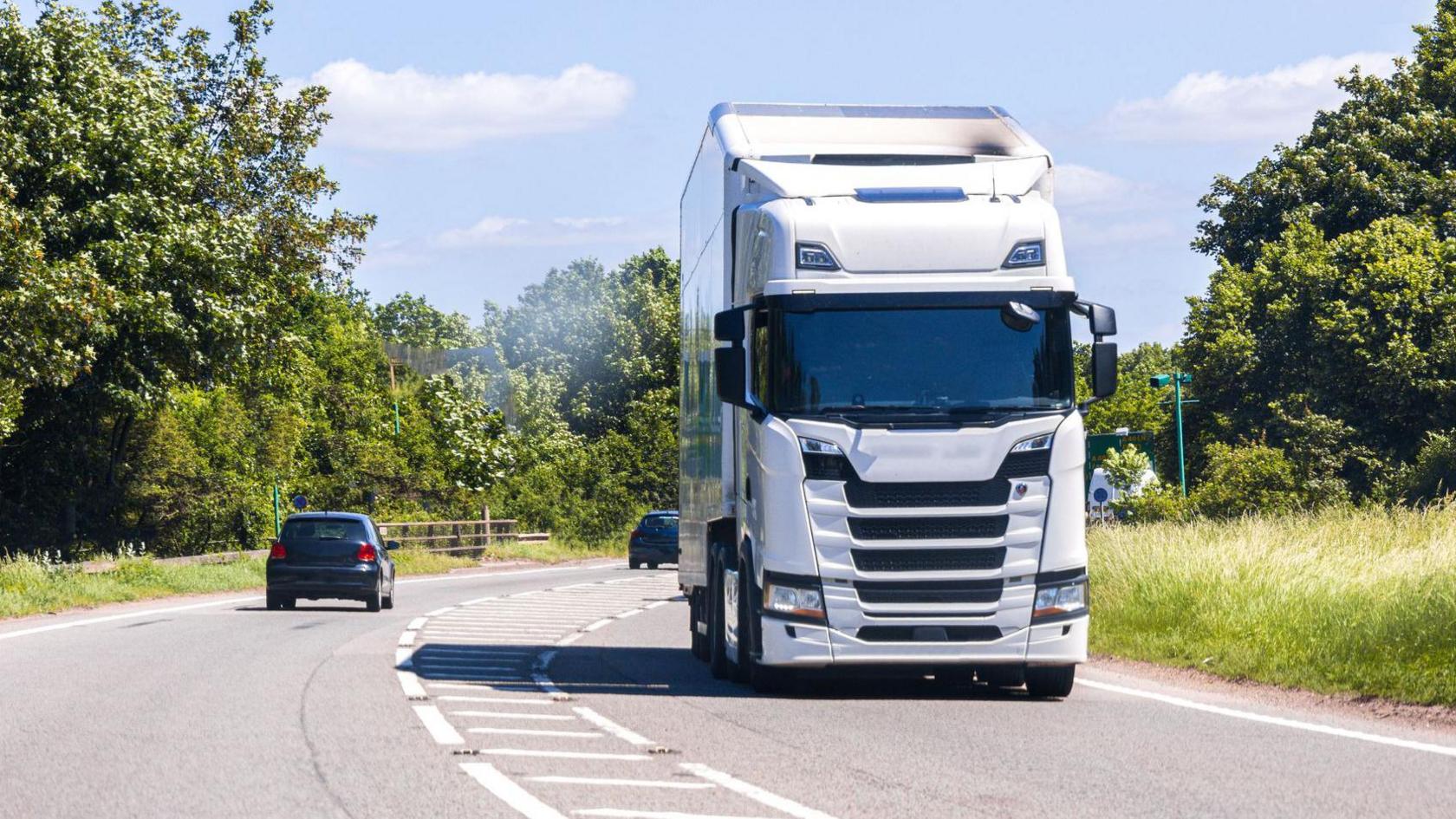 A white lorry passes two black cars going along a single carriageway road lined by trees