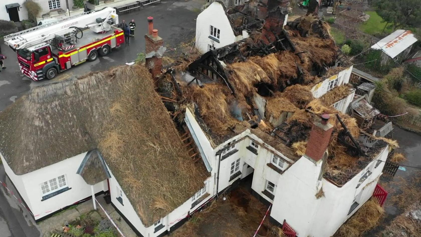 A drone shot of thatched cottages that have been damaged by a fire. The thatched roofs have been heavily burned. The homes are white painted and there is a fire engine parked on the road outside.