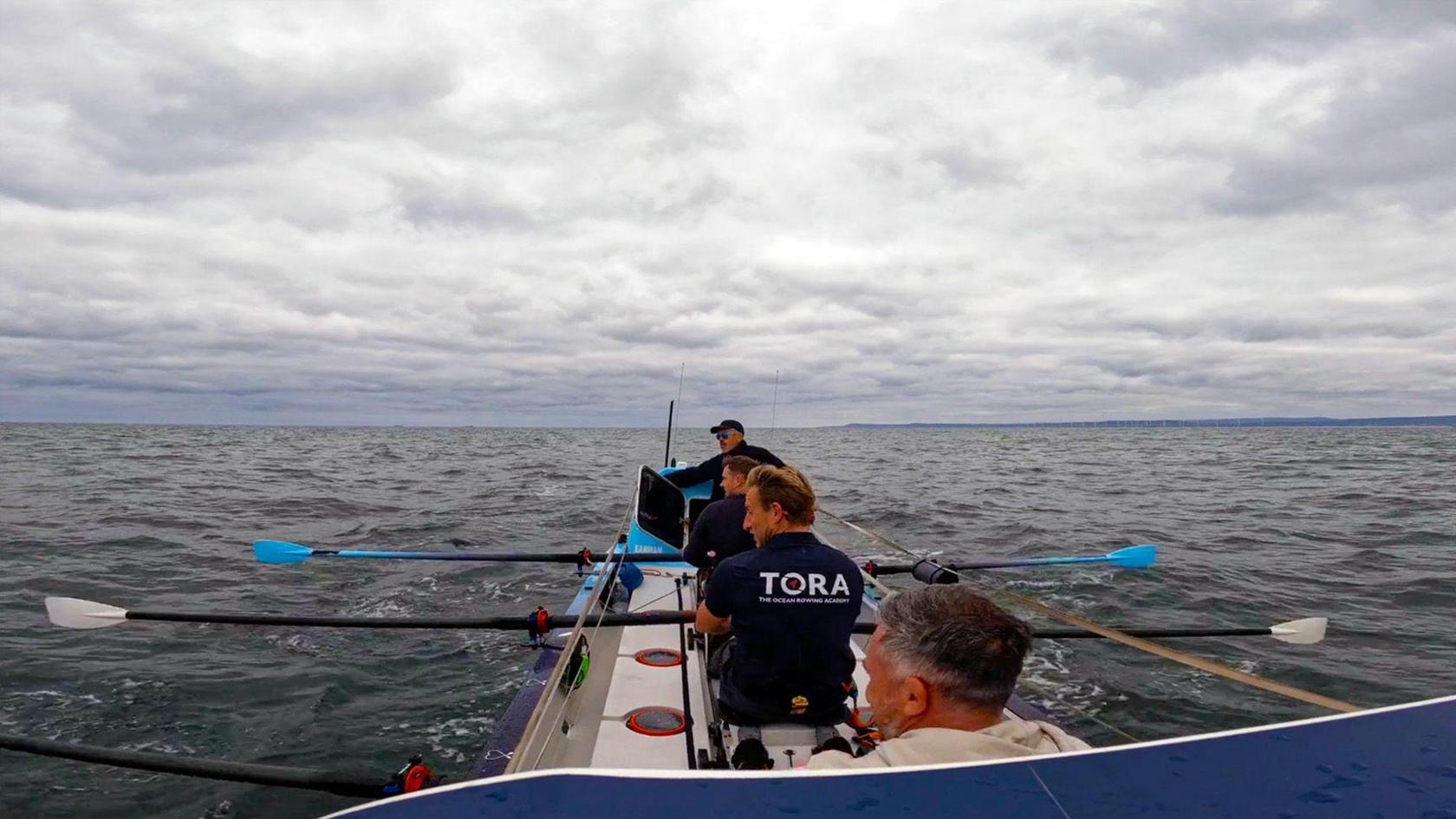 Four people in a rowing boar at sea, with shore visible on the horizon.