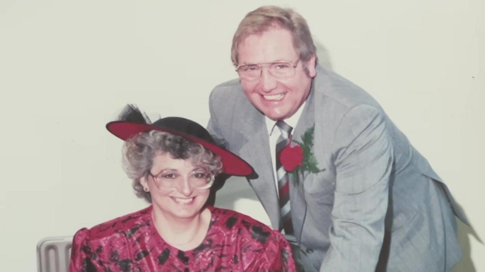 An older photo of David and Jenny Sexton, who are dressed up in smart clothes. Jenny is wearing a red and black hat and dress, while David is wearing a grey suit with a red rose in his lapel