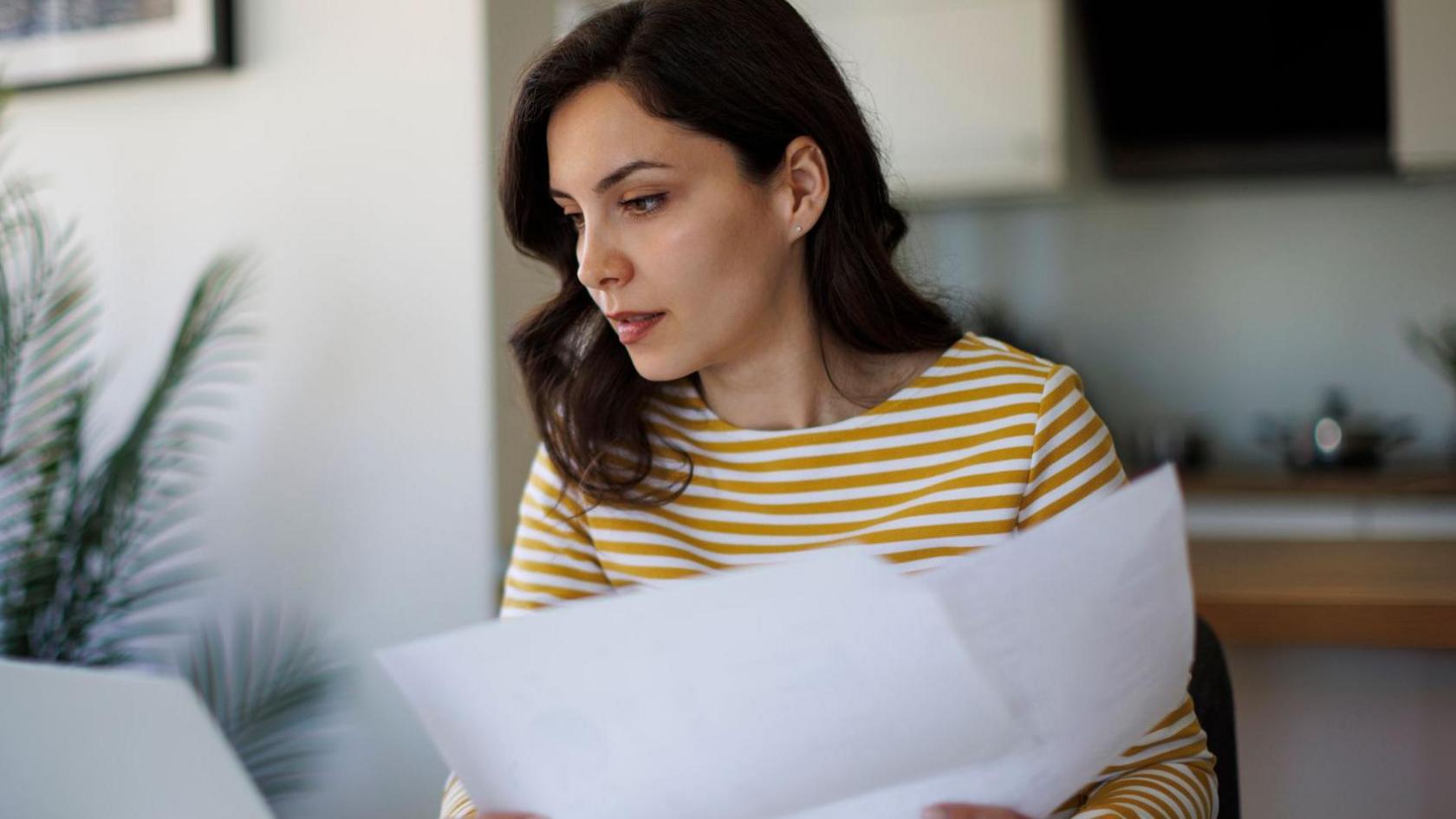 Woman looks at paperwork and a laptop at home with a pot plant behind her.