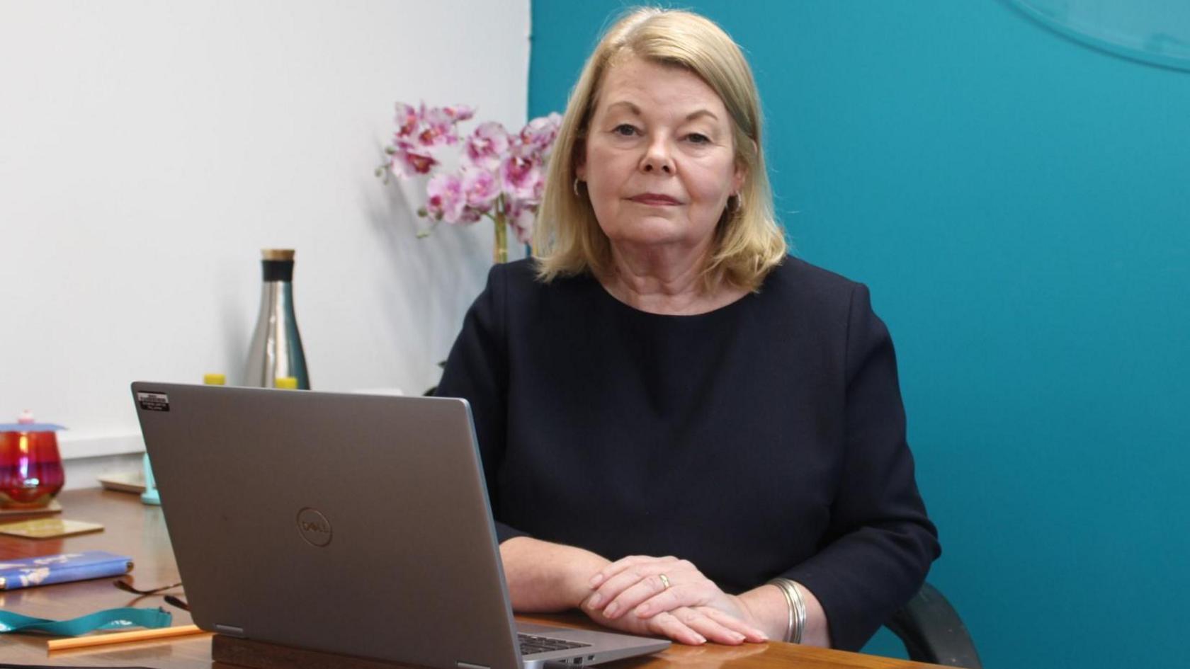 New Forest District Council leader Jill Cleary posing for the camera, sitting behind a desk with an open laptop. She has blonde shoulder length hair and is wearing a black, long-sleeve top.