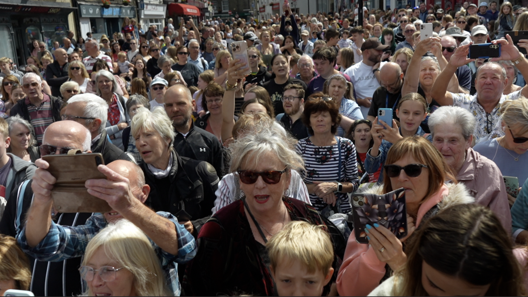 A crowd of hundreds of people fill a town centre road to watch an event, with shop fronts seen on the left side of the photo.