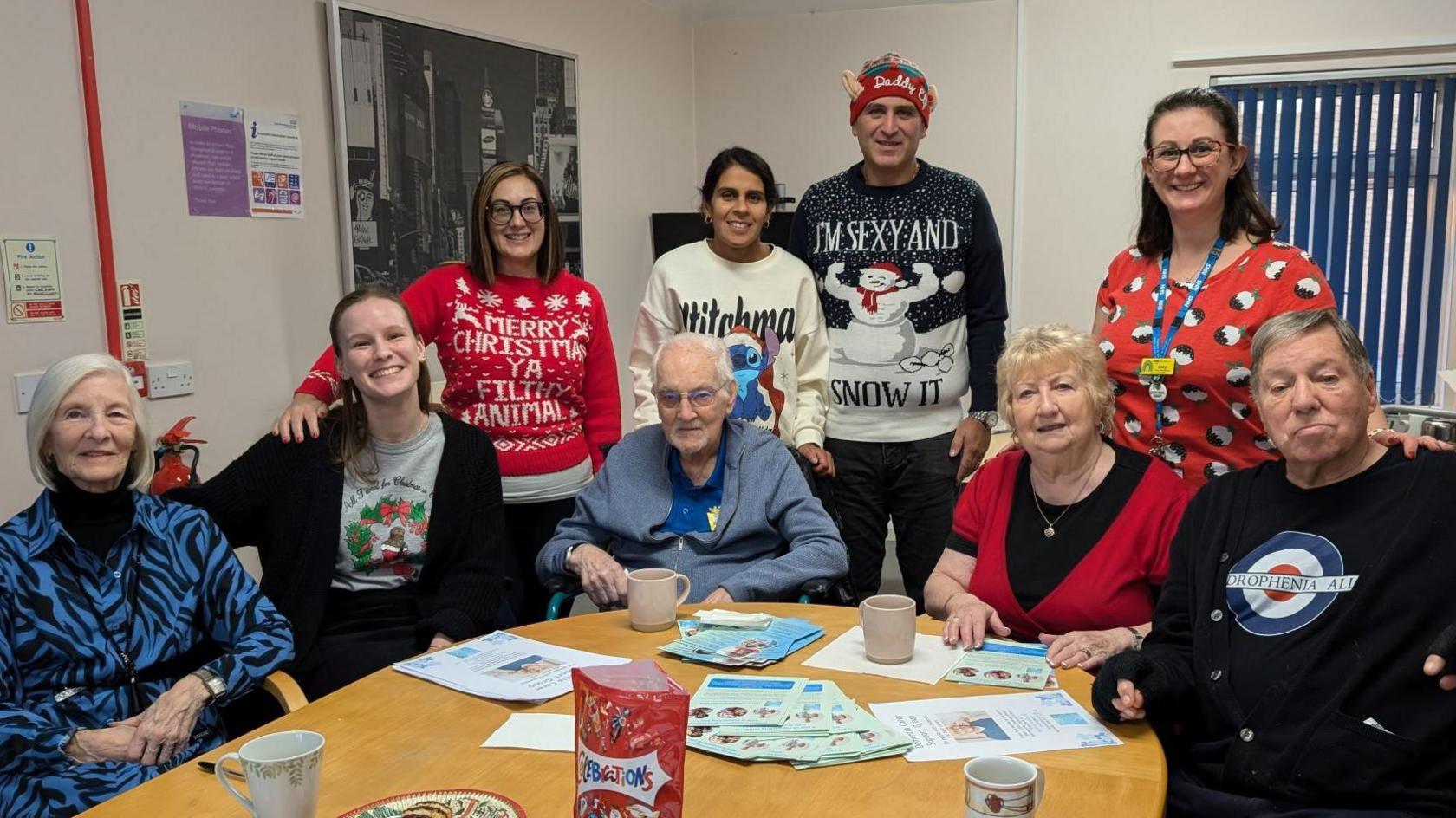 Six women and three men stood and sat around a table at the support group. They look happy and a few of them are wearing Christmas jumper.s