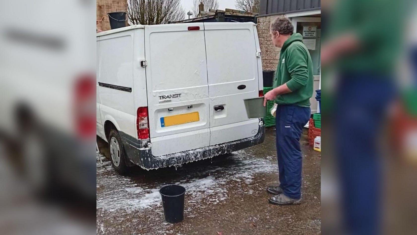 A man in a green hoodie and blue tracksuit bottoms cleaning his white van