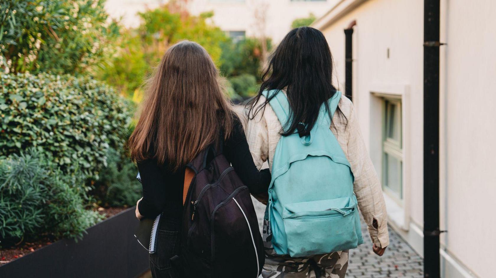 Two children walk away from the camera. The child on the left is wearing a black jumper and carrying a black rucksack, and has long brown hair. The child on the right has long dark hair and a blue rucksack, and is wearing a cream jacket. They are walking along a cobbled street with a building on their right and some bushes on their left.