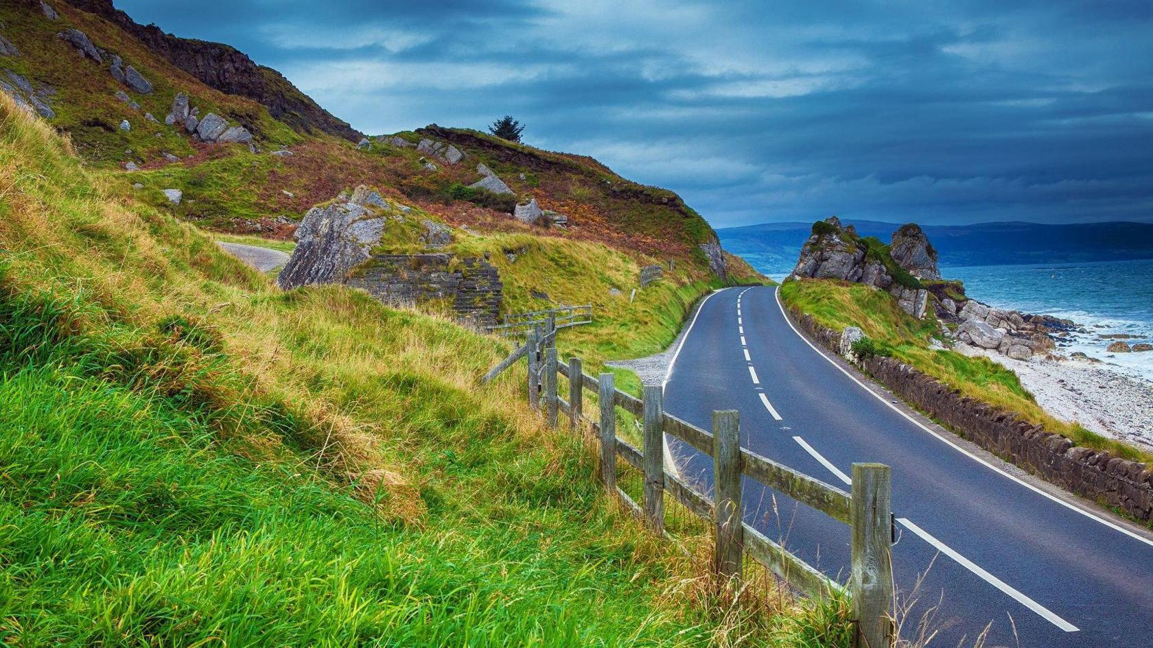 A road hugs the coastline in Northern Ireland, with the sea seen on one side, and rugged cliffs on the other.