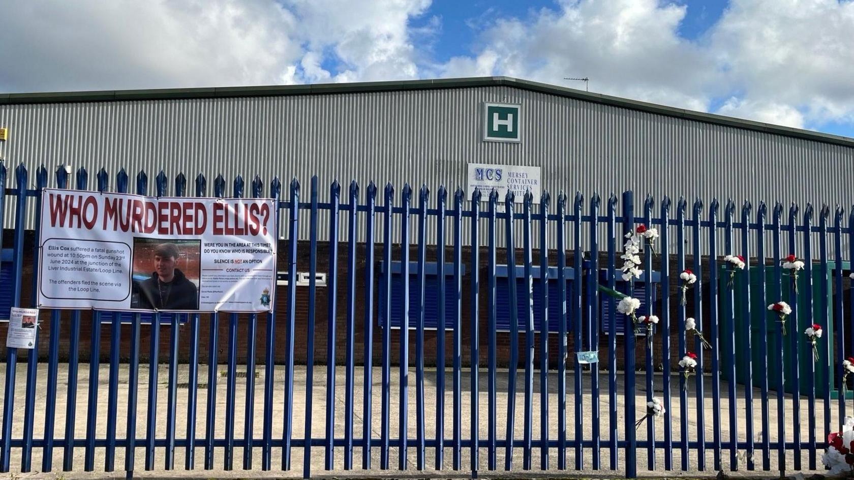 A banner with the words 'Who Murdered Ellis?' tied to a blue fence in front of a warehouse. There are also bunches of flowers tied to the fence