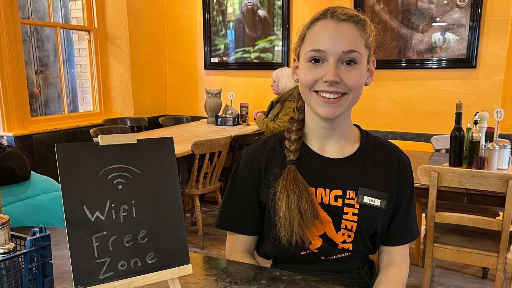 Lili smiling in a cafe by a small chalk board which reads 'wifi free zone'. She has long brown hair, tied into a plait, and is wearing a black t-shirt. Wooden tables can be seen in the background against a bright yellow wall. A customer is sitting at one of the tables. 