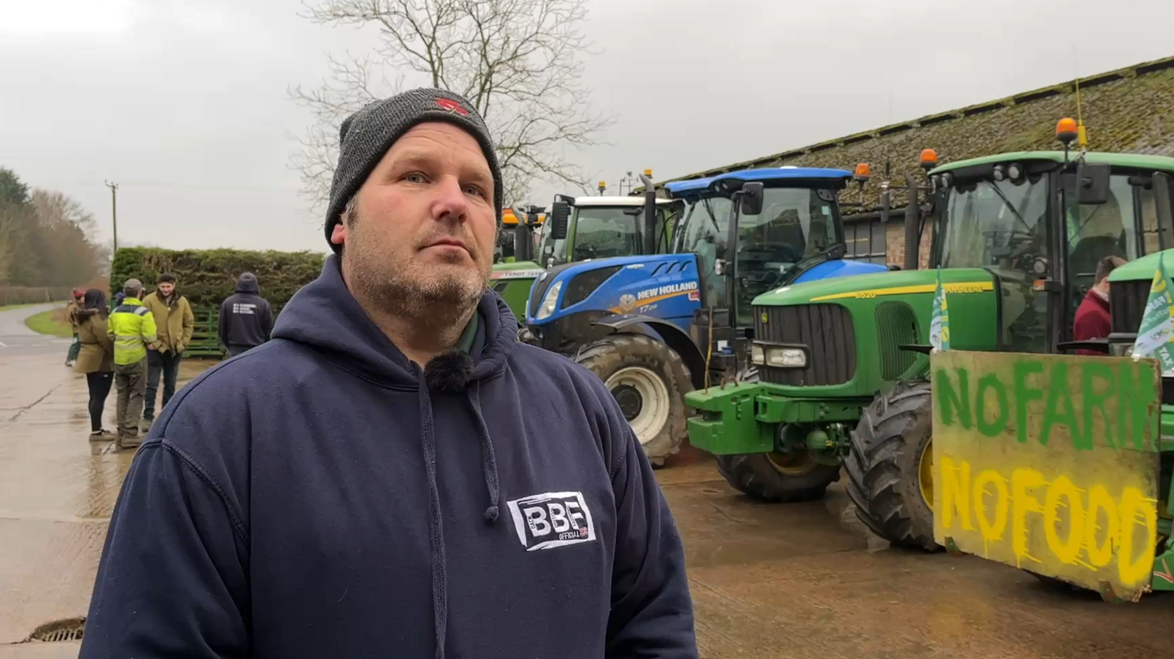 A farmer wearing a beanie and a navy hoodie stands in front of a line of tractors on a farm on a cloudy day. One of the tractors has a sign attached to it which has "No Farm, No Food" sprayed onto it with green and yellow writing.