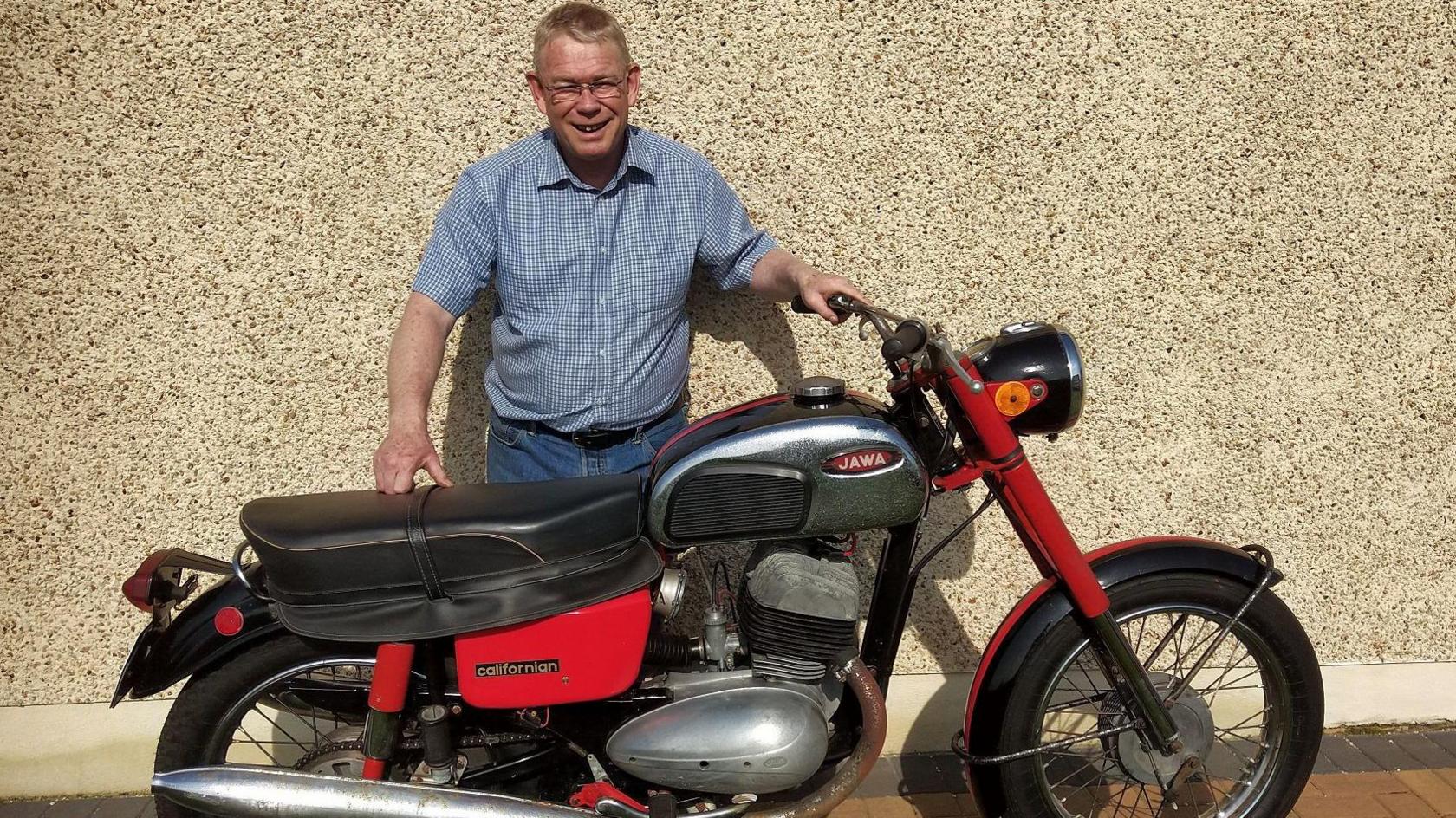 A man with short blonde hair, wearing glasses, a blue shirt and blue jeans, stands smiling at the camera behind a black and red motorcycle.