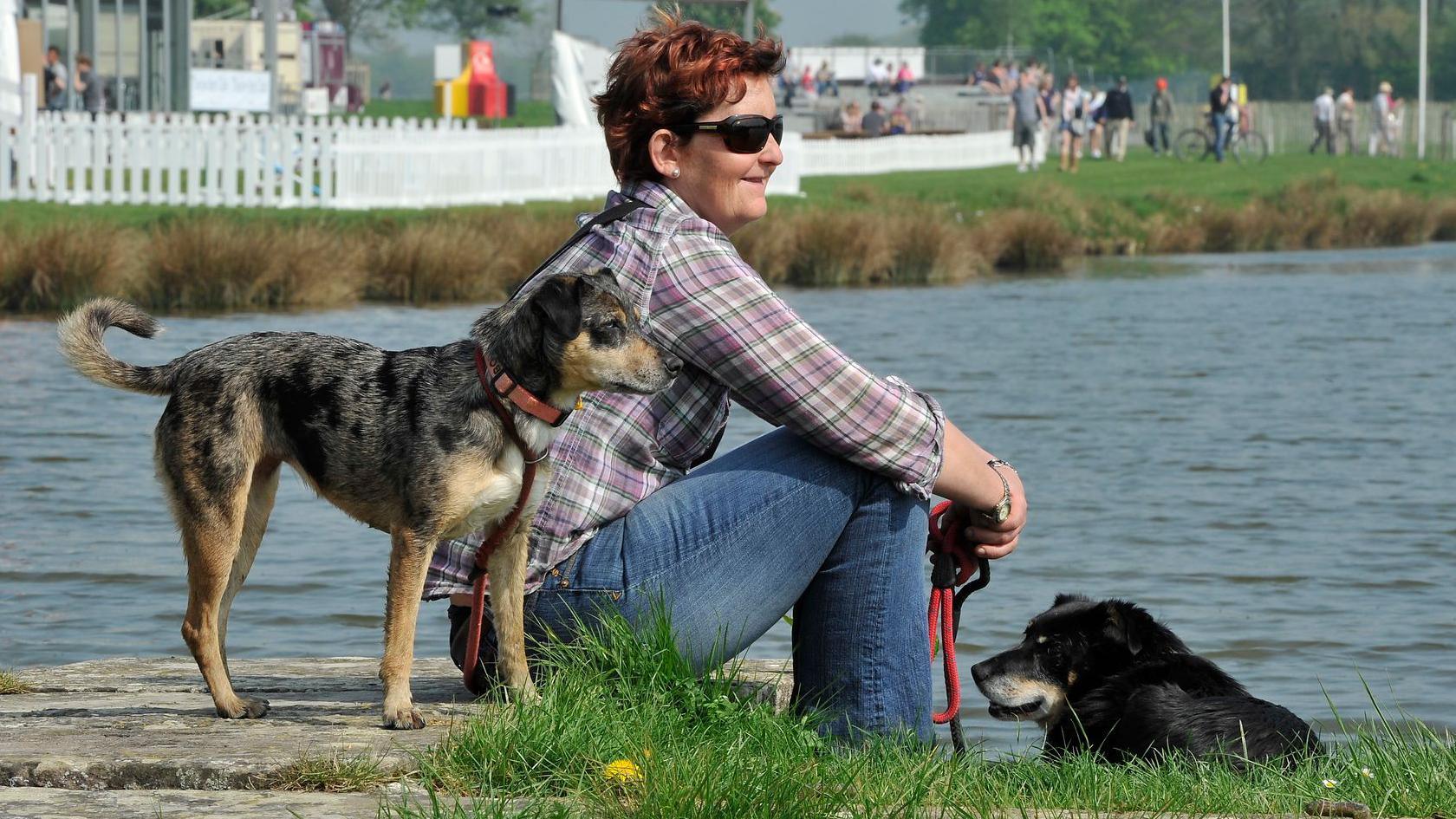 A woman sits with her two dogs at the lake in Badminton's cross country course.