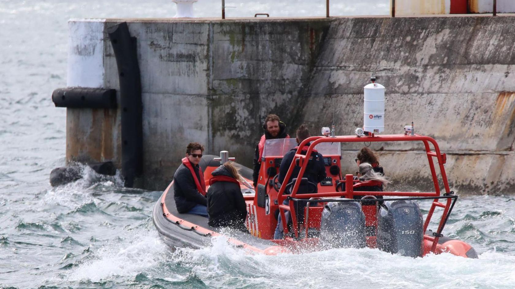 Tom Holland, wearing sunglasses and a red life jacket over a dark jacket, is sitting in an inflatable boat powered by two large outboard engines. There are at least five other people with him - three women and two men. The boat's engines are causing the water to foam as they head for the harbour entrance.