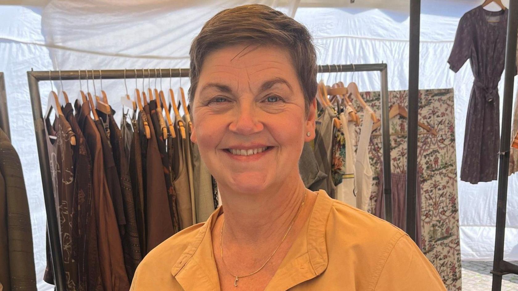 Ms Gregersen is smiling while standing at her stall. Behind her are metal clothing racks with clothes on wooden hangers. They are a range of designs in shades of brown, white cream and grey. She has short brown hair, blue eyes and is wearing a gold necklace with studded earrings. She is also wearing a yellow blouse.