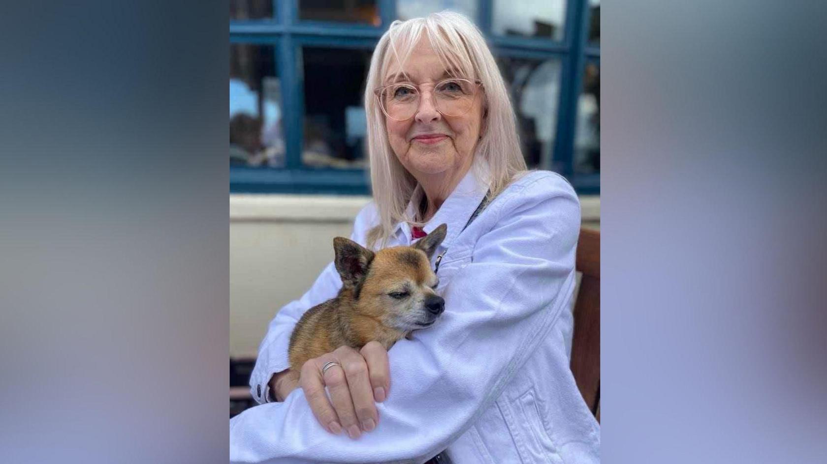 Jeanette Walker, with white hair and pink glasses, wearing a lilac coloured jacket, holds a small dog while smiling at the camera