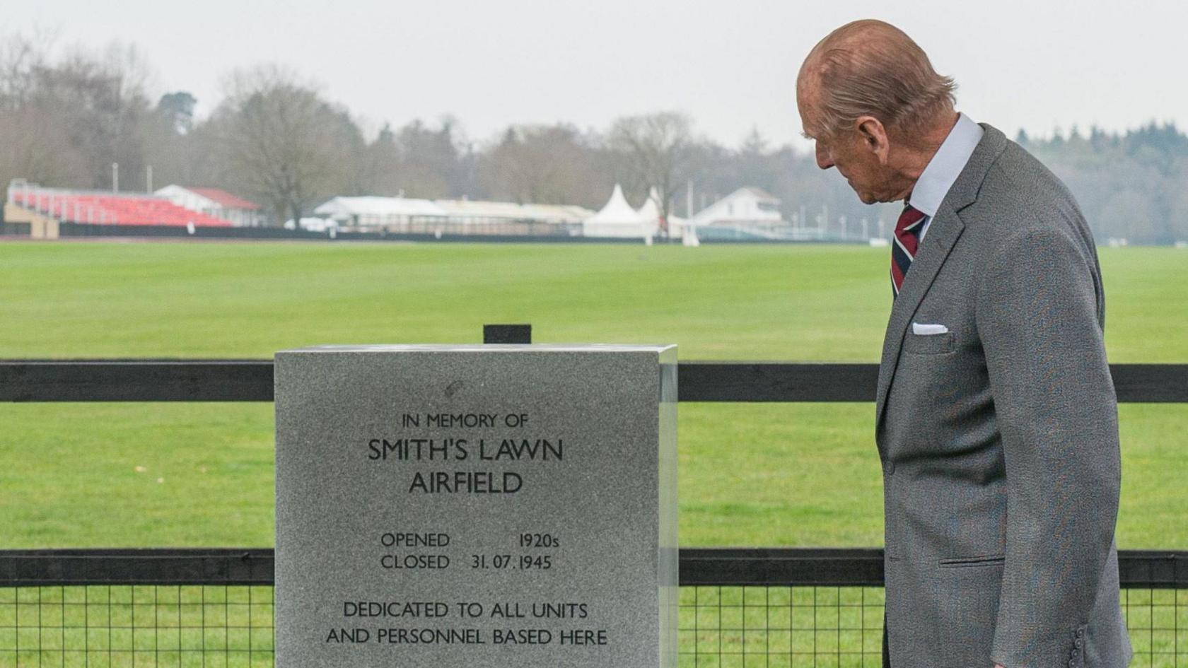 The Duke of Edinburgh is wearing a grey suit and grey shirt, with a red, white and black tie. He is looking at a grey memorial block which reads "In Memory of Smith's Lawn Airfield". Behind him is what used to be the airfield site, which is now a polo field and club.